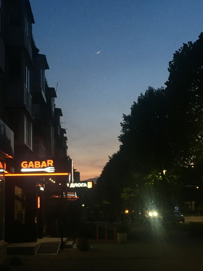 A nighttime street scene in Lutsk, Ukraine, under a dark blue sky with a faint crescent moon. On the left, a multi-story building with a brightly lit orange sign that reads "GABAR" is visible. Across the street, silhouetted trees line the right side of the frame. Streetlights illuminate the sidewalk and road, and the headlights of a car are visible in the distance, creating bright streaks of light. Another illuminated sign with Cyrillic text is partially visible further down the street. The overall atmosphere is calm and depicts a typical urban evening.