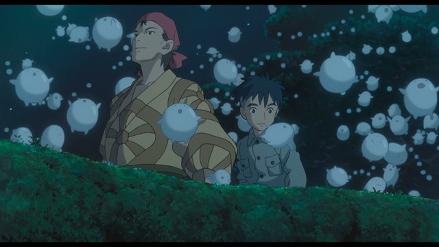 Still from The Boy And The Heron that depicts Mahito, a shaggy-haired boy in grey, standing with Kiriko, a woman waring a yellow yukata with a wheel motif and a red bandana. They're both looking in wonder at mysterious bubble-like creatures that are rising up from the greenery around them.