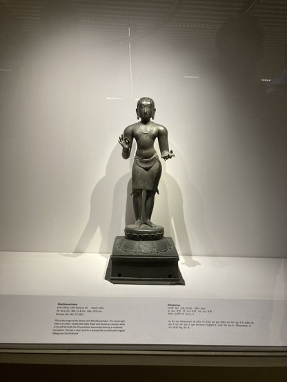 Shaiva saint Manikkavachakar. The raised right hand in china -mudra (the index finger and thumb are joined); while in the left he holds the Tiruvachkam manuscript bearing a mutilated inscription. The hair is short and he is dressed like a monk with ringlets falling over the forehead.