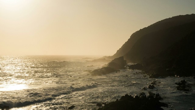 Golden hour sunlight illuminates a rocky coastline with waves crashing against the shore.