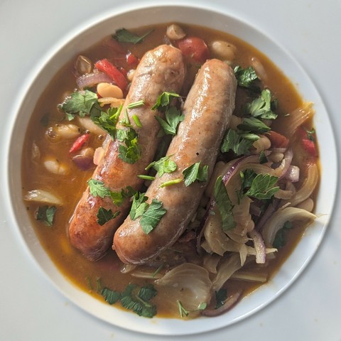 White bowl, two large sausages sitting in a brothy stew with bits of fennel, beans and peppers, coarsely chopped parsley over the top