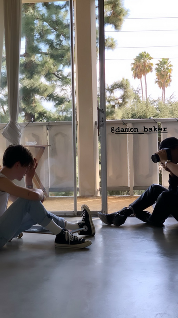 Two young men sit on the floor, one photographing the other. The model wears a white T-shirt, blue jeans and black Converse boots with white socks, and there’s a skateboard next to him on the floor. The photographer wears dark blue jeans, a black tshirt and hat, and his face is obscured by his camera.