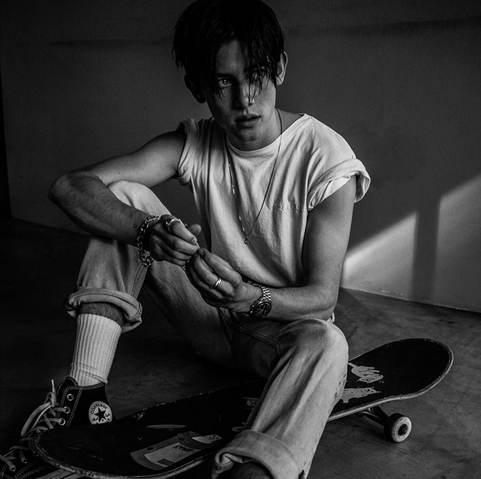 Black and white photo of a young man sitting on the floor, with a skateboard next to him, and looking into the distance. The sleeves of his white tshirt are rolled up as are his jeans, showing his white socks. He’s wearing a necklace, bracelet, rings and watch.