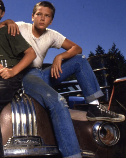 River Phoenix as Chris Chambers in the film Stand By Me, sitting on the hood of a car. He’s wearing a white tshirt, blue jeans with the ends rolled up, white socks and black hightop sneakers like Converse boots.