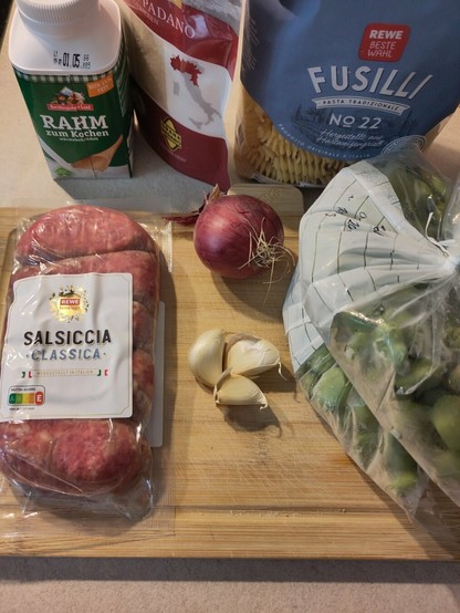 Die Zutaten für das Gericht, darunter die Salsiccia, Zwiebel, Knoblauchzehen, Puffbohnen, Nudeln, gerienener Hartkäse und Kochsahne.
