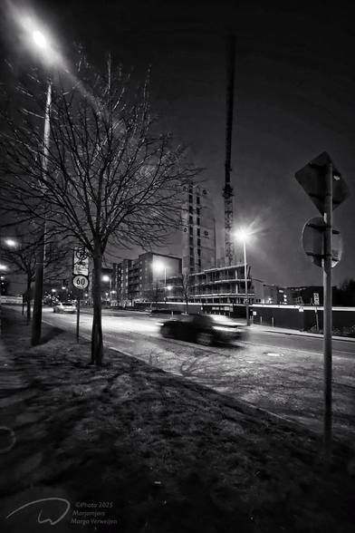 Very early in the morning, it is still dark, street lamps give light, trees along the road make their shadow on the road surface. A car drives quickly past in the now almost empty street before the traffic jam begins. On the other side of the road a new apartment complex rises. A crane towers high in the air.