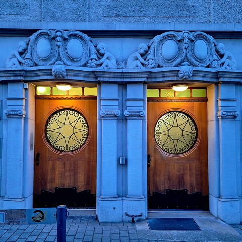 A picture shows twin wooden doors framed by stone columns and topped with stone shields and cornucopias. Four sculpted children reach for fruit spilling from the cornucopias. Circular windows with star-shaped grilles are set into the doors.
