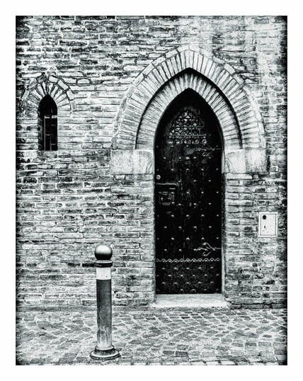 A black and white photo of the wall of a building from a street. On the left hand side there is a small arched window. On the right hand side an vaulted door with studs on it. in the foreground between the window and door is a small bollard at street level.