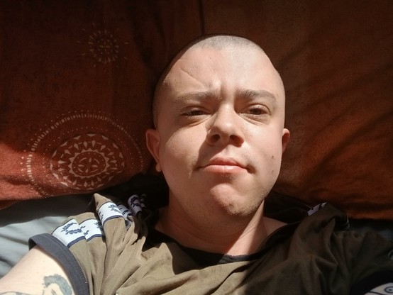 A white trans mask wearing a green top lying in bed. He has a shaved head and the sun is on his face