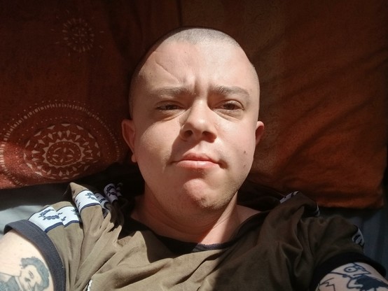 A white trans mask wearing a green top lying in bed. He has a shaved head and the sun is on his face