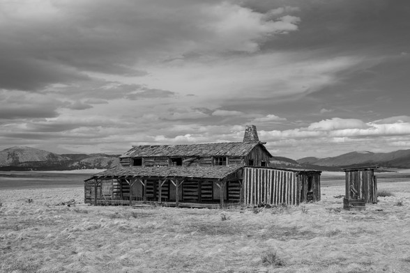 A weathered log cabin and a small outhouse stand in an open grassy field under a wide sky, with distant mountains in the background. The photo is black and white, emphasizing texture and contrast. This is a preserved movie set in Valles Caldera, New Mexico.