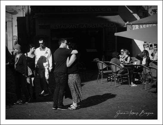The image captures a vibrant street scene depicted in black and white, bringing a timeless quality to the moment. In the foreground, a couple is affectionately embracing, creating a focal point of intimacy. The man is dressed in a simple black t-shirt and jeans, while the woman wears patterned pants, reflecting a casual yet stylish look.
Surrounding them, several people are engaged in various activities. To the left, a group of friends walks closely together, their expressions showing a mix of joy and casual conversation. In the background, the ambiance of the location comes alive with a cozy restaurant sign reading “Restaurant La Pastachuca,” hinting at a lively outdoor dining scene.
A few patrons sit at tables under large umbrellas, absorbed in their conversations or enjoying their meals. The sunlight casts soft shadows, enhancing the depth and texture of the scene. This juxtaposition of companionship and everyday life highlights themes of connection and community, inviting the viewer to reflect on the relationships that fill urban spaces with warmth.
The artist’s choice to use black-and-white photography emphasizes emotional resonance over color, allowing for an exploration of light, shadow, and form that evokes nostalgia and simplicity.
