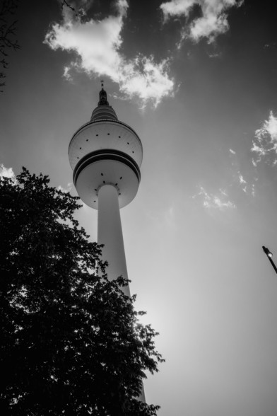 The black-and-white photo captures a dramatic upward view of the Heinrich-Hertz-Turm (Hamburg TV tower) silhouetted against the sky, with leafy trees and clouds adding depth and contrast to the composition.