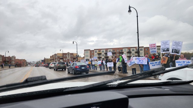 The image shows a protest scene viewed from inside a vehicle. In the foreground, the dashboard and windshield wipers are visible, with raindrops on the windshield. The scene is set on a street with a group of people standing on the sidewalk, holding signs. The protesters are wearing various types of clothing, including jackets and hoodies, suggesting a cool or rainy day. The signs they are holding are not fully legible, but one sign reads "HAND OFF." The background features a multi-story brick apartment building with balconies, and several cars are parked or moving on the street. The sky is overcast, and the overall atmosphere appears to be that of a peaceful demonstration.