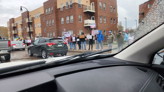 The image depicts a street scene with a group of people standing on the sidewalk, holding signs during a protest. The protest appears to be taking place on a cloudy day, with overcast skies and wet road surfaces, suggesting recent rain. The participants are holding various signs with messages such as "Rule of Law Matters." The signs are predominantly white with blue and red text, and some include images. The protesters are standing in a line, facing the road, with several cars stopped behind them, indicating a traffic stoppage. The background features a row of multi-story buildings with a brick facade, typical of urban residential architecture. The scene is viewed from inside a vehicle, as indicated by the visible dashboard and windshield wipers in the foreground.