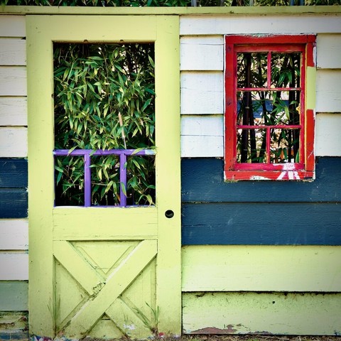 A bright-safety-yellow, glassless wooden door holds a portion of the viewing window's purple-painted pane frames within, as it stands next to a bright red, glassless wooden window, whose housing also holds a portion of its pane frames within, both of which share a wall of wooden slats (from colors white, down to blue, down to safety-yellow), and both of which lead into a grove of bamboo trees beyond their respective thresholds.