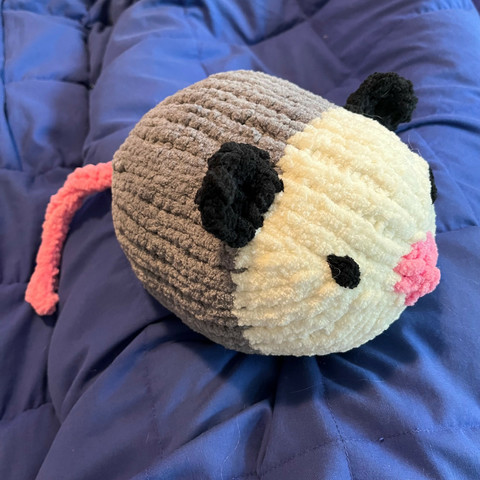 A large knitted opossum made of fuzzy chenille yarn, maybe 8 inches across, with a blunt pink nose and embroidered black eyes