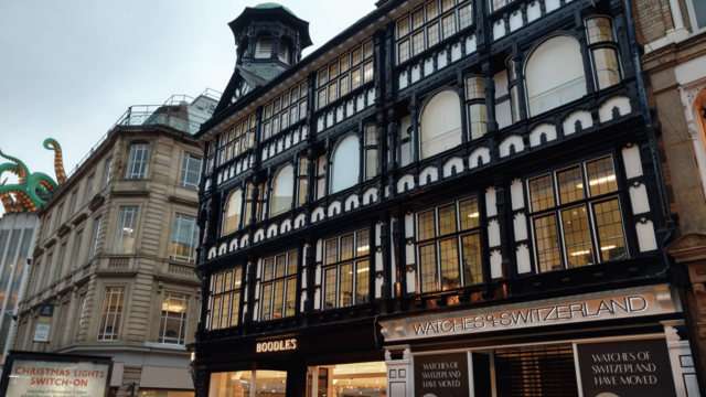 Ein prachtvolles, sehr breites Fachwerkhaus an einer schmalen Straße. Die deutlich sichtbaren Holzbalen sind schwarz lackiert, die Flächen sind weiß oder befenstert. Oben an der Hausecke sitzt ein Türmchen mit Giebel. Die Fassada trägt Werbung und Firmenschilder von Uhren- und Luxusgeschäften. etwa Watches of Switzerland und Boodles. Am linken Rand des Bildes schauen oberhalb der Fassade lange, grün-geölbe Tentakel aus dem übernächsten Haus, welche sich in den Himmel zu strecken scheinen.