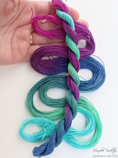 Skeins of thread for lacemaking that have been dyed by hand in colors shading from magenta, purple, blue, and aqua displayed on a hand to show off the colors.