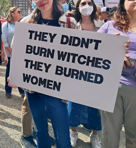 A woman holds a protest sign on a street in Philadelphia, as she stands with other protesters.  The sign reads, "They didn't burn witches, they burned women."