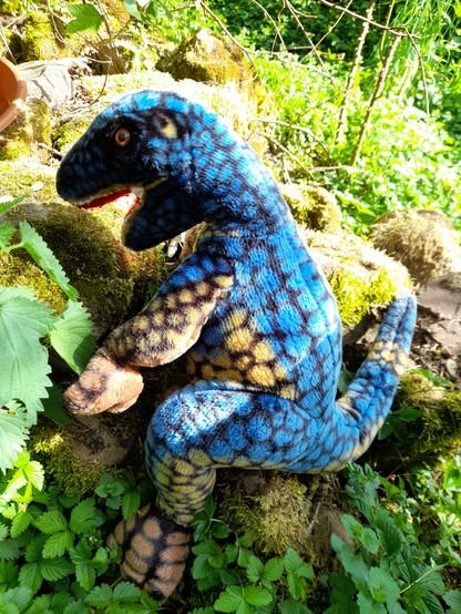 A rather big lezard-headed blue plush dinosaur climbin on some sandstones between wild herbs.