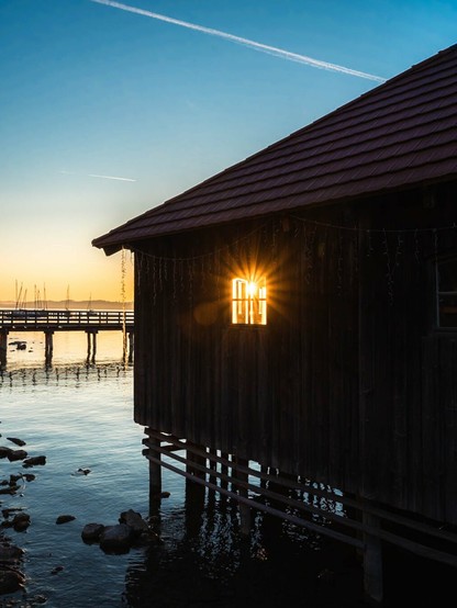 This image shows a peaceful lakeside scene at sunset. In the foreground is a wooden boathouse or cabin built on stilts over water. The structure has dark wooden walls and a brown shingled roof. A window on the side of the building is capturing the setting sun, creating a dramatic starburst effect as the sunlight shines directly through the glass.

The boathouse stands in calm water where small rocks are visible beneath the surface. To the left, you can see part of a wooden pier or dock extending into the water. In the background, there are what appear to be sailboat masts visible near the shoreline.

The sky is a beautiful gradient, transitioning from deep blue at the top to golden yellow near the horizon where the sun is setting. A single contrail from an airplane cuts across the blue sky. The overall atmosphere is serene and golden, capturing that magical moment when day transitions to evening over a tranquil body of water.
