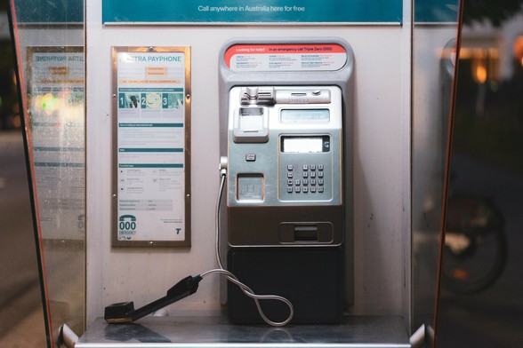 The image shows a public payphone. It includes a handset, dial pad, and display screen, with instructions visible on the side. It's installed in a booth, and there's a sign indicating it's for making calls, possibly in Australia given the wording at the top.