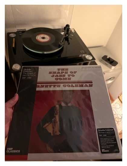 Auf einem schwarzen Plattenspieler läuft eine schwarze Schallplatte. Davor halte ich das Cover ins Bild. Weißes Cover, Foto von Saxophonist Ornette Coleman vor rotem Hintergrund, Titel und Künstler in roter Schrift.