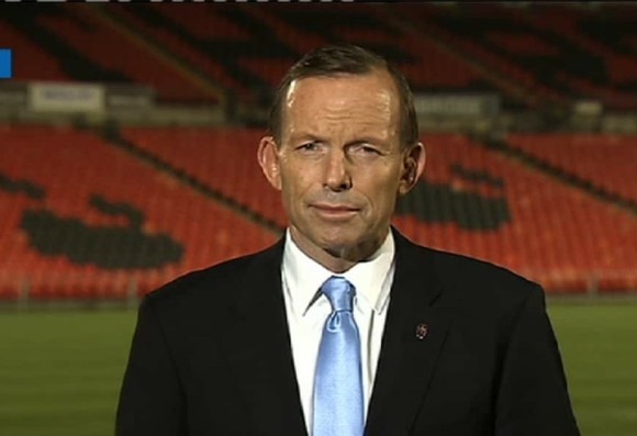 Tony Abbott stands in front of empty stadium seats, looking serious. The background features orange seats with dark patterns.