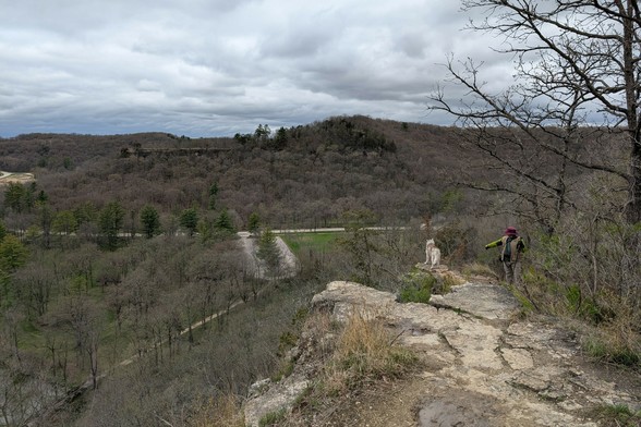 Jeff continues to photograph Zeena on the bluff, while the photographer has passed and is looking down from an even higher overlook on the edge of the bluff. From here, you can see the valley below, and bluffs of similar height on the far side. A road cuts through the valley, and a park parking lot can be seen between trees below. The flat, textured cloud layer undulates over the far hills.
