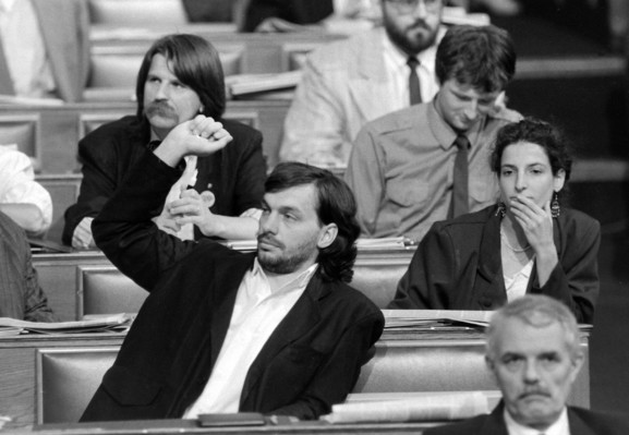 A black and white photo of several people sitting at desks in Parliament. In the foreground is a young bearded man, Viktor Orbán, with his arm half-raised, as if indicting he wants to say something.  Behind him is another man with longish hair and very large moustache. There are also other MPs visible in the photo.