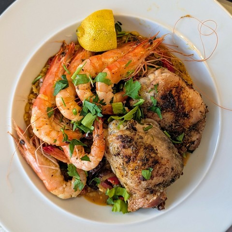 White bowl, head on prawns and chicken thighs top deep orange/red rice, coarse strewn parsley and a lemon wedge on top