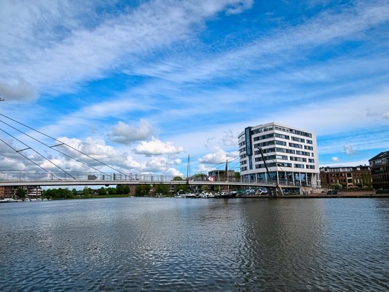 In the foreground, the water of a harbour. A curved bridge spans the water. A box-shaped white tower block can be seen on the other bank. The sky is mainly blue with white streaks of cloud and individual fluffy clouds.