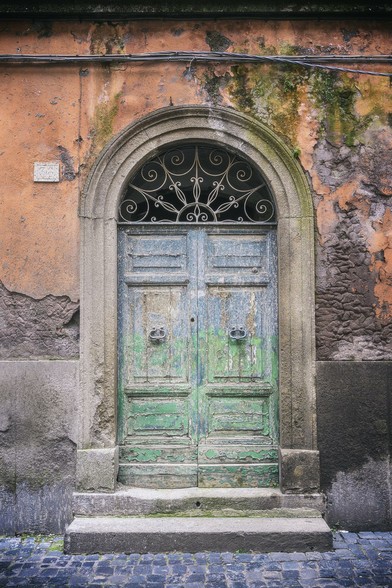 In Viterbo Italy, an old wooden door with peeling green and blue paint sits in a stone archway, surrounded by a weathered orange and gray concrete wall. Intricate ironwork above the door adds a touch of elegance to the rustic facade.