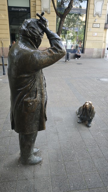 A bronze statue of the star of the TV crime series lieutenant Columbo in a typical pose, scratching his head with the end of a cigar in his fingers. Seen from the side. In front of him an occasional character, a dog, I guess the basset breed, sitting and looking up to him. Seen from the front. The whole scene is on the street.
