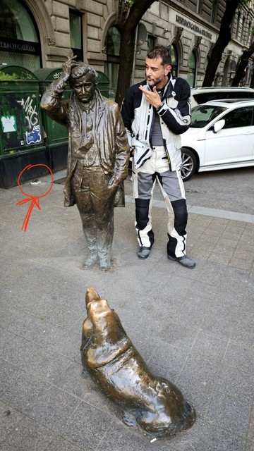 The same statues, this time Columbo seen from the front, the dog from the back. Next to the lieutenant stands a person (in white/black textile motocycle pants and jacket, making a thinking pose to like they are trying to solve the case together. Behind them on the pavement is a small spot, circled in red with an arrow added to the photo.