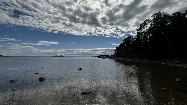 View over the water with some cloud coverage, trees in the right, small isalnds, and some stones sticking up out of the shallow water.