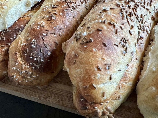 Auf einem dunklen Holztisch liegt ein Holzbrett. Darauf liegen zahlreiche frisch gebackene Brötchen mit rustikalem Ausbund.