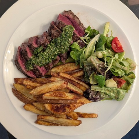 White plate, thick slices of griddled rare venison with salsa verde on top, pile of skin-on chips, dressed salad