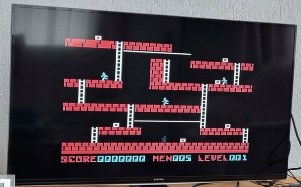 The first level of the game Lode Runner on a Samsung tv. The level is made up of different red platforms with white ladders connecting the platforms. A dark person-shaped sprite [player character] is seen on the bottom, three white [enemy] characters are on higher platforms. Spread out over the level are several white-and-read squares. Text at the bottom shows the current level info: "Score0000000 Men005 Level001