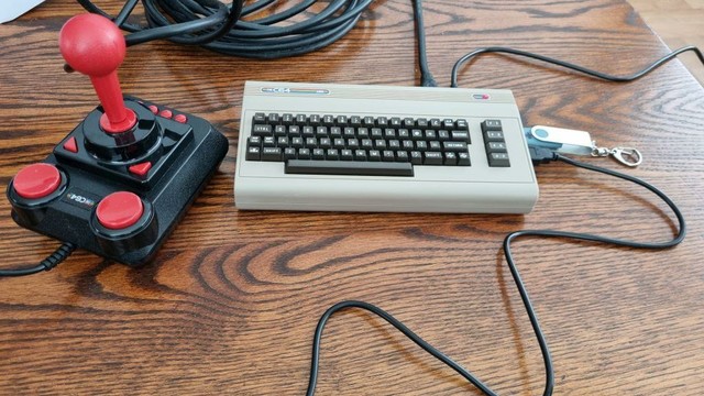 TheC64 Mini computer, a mini replica of the Commodore 64 breadbox computer, next to a full size joystick similar to the Competition Pro. A cable runs from the right side of the computer to joystick, which is placed to the left of the computer. A USB stick is plugged into the right side as well. A cable runs from the back right of the computer to something out of view to the right of the picture [power supply]. Another, coiled, cable runs from the back right to something out of view to the top of the picture [HDMI cable to tv]

Colors: the C64 is beige with black an grey keys like the original, the joystick is black with a red grip. The USB stick is blue with a metal keychain part at one end. All is placed on a wooden background of different shades of brown.
