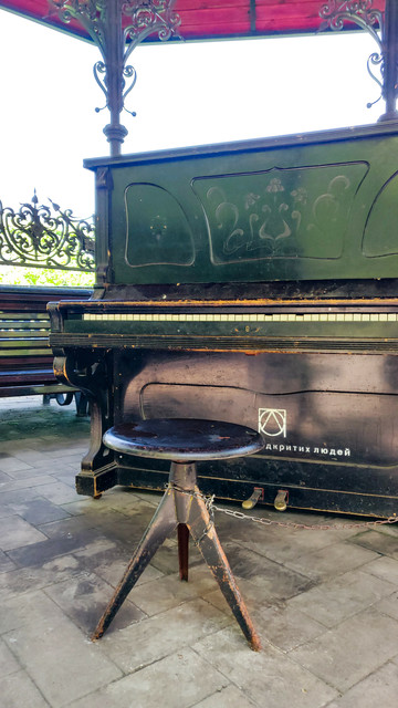 Unter einem Pavillon mit tiefrotem Dach und verschnörkelten gusseisernen Streben steht ein altes mattschwarzes Piano. Ein runder, drehbarer Klavierhocker ist daran angekettet. Oberhalb der Klavier-Pedale sind ein Logo aus geometrisch ineinander verschachtelten Formen und der teilweise vom Hocker verdeckte Schriftzug покоління відкритих людей erkennbar.