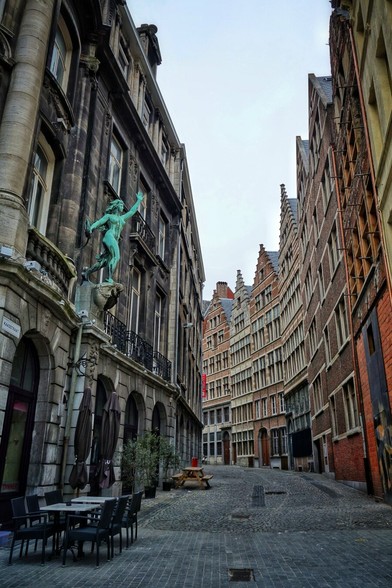 A ground-level, slightly low-angle shot looks down a narrow, cobbled street lined with tall, ornate buildings. The building on the left is dark grey stone with arched ground-floor openings, some of which appear to be part of a restaurant with outdoor seating consisting of black tables and chairs. A striking, verdigris-coloured statue of a figure reaching upwards is mounted on the corner of this building, above a decorative balcony. The buildings on the right are predominantly red brick with varying architectural styles, including stepped gables. The street curves slightly into the distance, revealing more of these historic-looking buildings under an overcast sky.