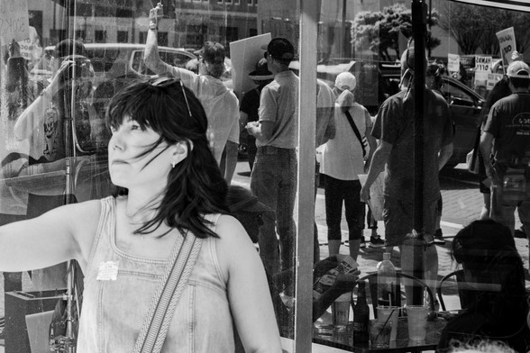A woman standing in front of a plate glass window taking photos with her phone of the "Hands Off" protests happening on the street, as can be seen in the reflections from the window.  I focused on the reflections.