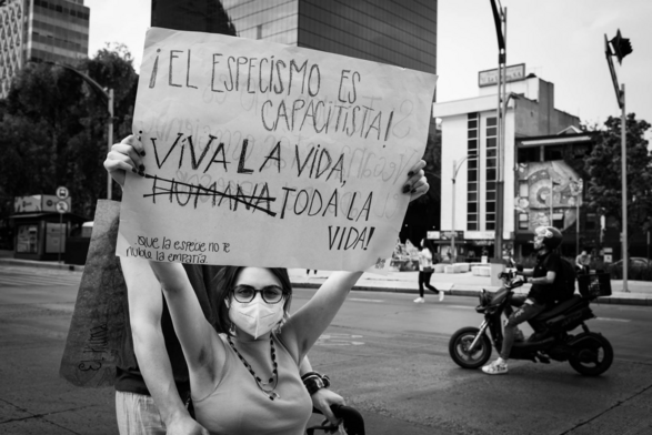 Fotografía en blanco y negro. Una activista con gafas, sentada en una silla de ruedas, sostiene un cartel blanco con letras negras que dice: ¡EL ESPECISMO ES CAPACITISTA! ¡VIVA TODA LA VIDA HUMANA (la palabra humanan está taschada) TODA LA VIDA! Que la especie no nuble tu empatía. Al fondo de la imagen a la derecha se ve pasando un motociclista y en el fondo de la imagen se pueden observar edificios, una lámpara y un semáforo. © Sofía Dumat|CLAMOR DE LIBERTAD