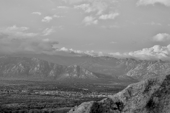 View of a valley from some heights with the village of Cerceda at the bottom and La Maliciosa mountain behind. It is a picture in black and white with some sky and clouds on top.