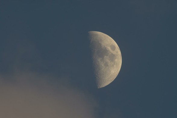 Der zunehmende Halbmond am blauen Tageshimmel, von unten links schiebt sich eine Wolke bis kurz vor dem Mond ins Bild