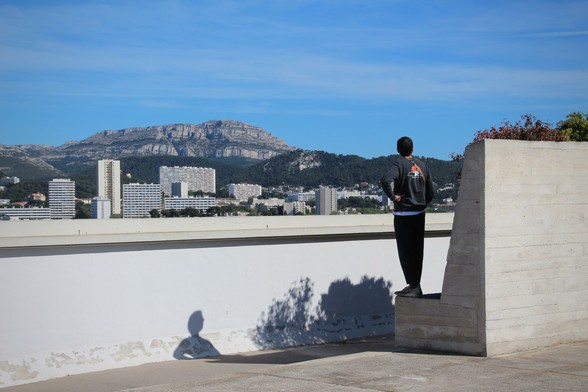 Ein junger Mann steht auf einer Betonkonstruktion und schaut über eine niedrige Mauer in die Weite. Man sieht Hochhäuser und einen markanten Berg mit flachem Gipfel. Blauer Himmel.