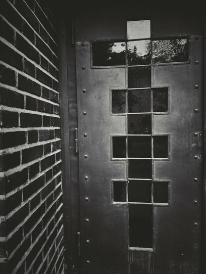 The portrait-orientated black and white image shows a close-up view of a brick wall and a metal door. The wall is on the left side of the image and is constructed of rectangular bricks laid in a pattern. The metal door is on the right side of the image. It is made of solid material with visible rivets or fasteners. The door has a pattern of multiple small, rectangular glass panes. The panes are arranged in a grid-like structure that forms a cross shape.