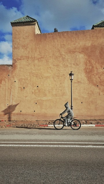 The portrait-orientated image shows a person on a bicycle riding past a wall. The person is wearing a traditional Moroccan hooded garment jelaba. There is a street lamp visible. The wall is made of light brown material and has a green tiled roof section. The ground is paved with asphalt and includes white lines marking lanes. There is a striped red line painted on the ground near the base of the wall, marking the beginning of the pavement. The sky is partly cloudy. Shadows are cast on the wall and the ground.